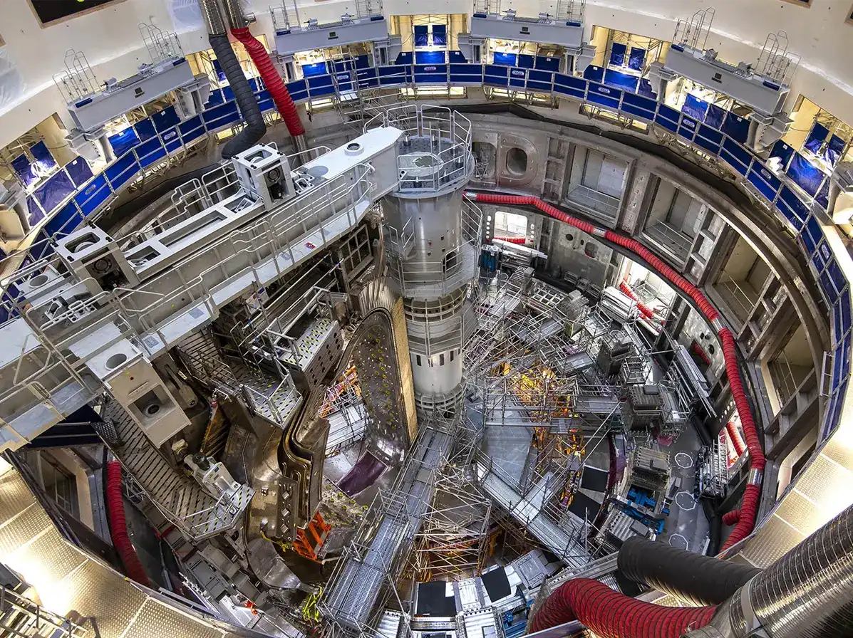 Hala de asamblare a tokamak-ului ITER — vedere de sus în etapa de construcție, Cadarache, Franța Vedere aeriană fisheye a interiorului halei tokamak ITER în etapa de asamblare, cu platforma circulară, structuri metalice, eșafodaj și conducte de ventilație roșii — șantierul de construcție al celui mai mare reactor de fuziune nucleară din lume